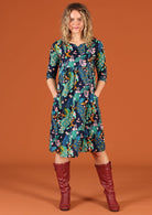 Woman models a knee length cotton dress with 3/4 sleeves in a peacock print, with hands in pockets