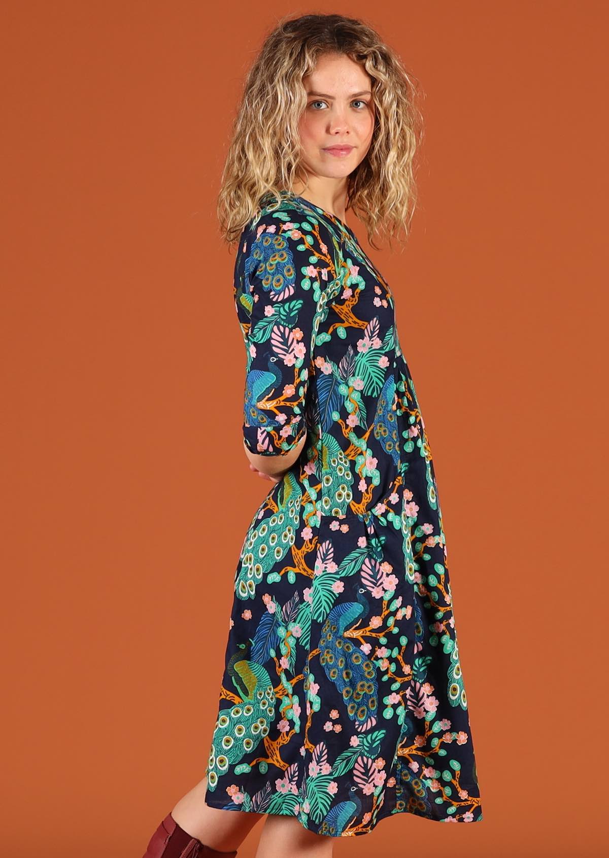 Woman standing side on wearing cotton knee length dress in a peacock print, with arms behind her back