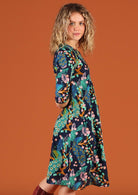 Woman standing side on wearing cotton knee length dress in a peacock print, with arms behind her back