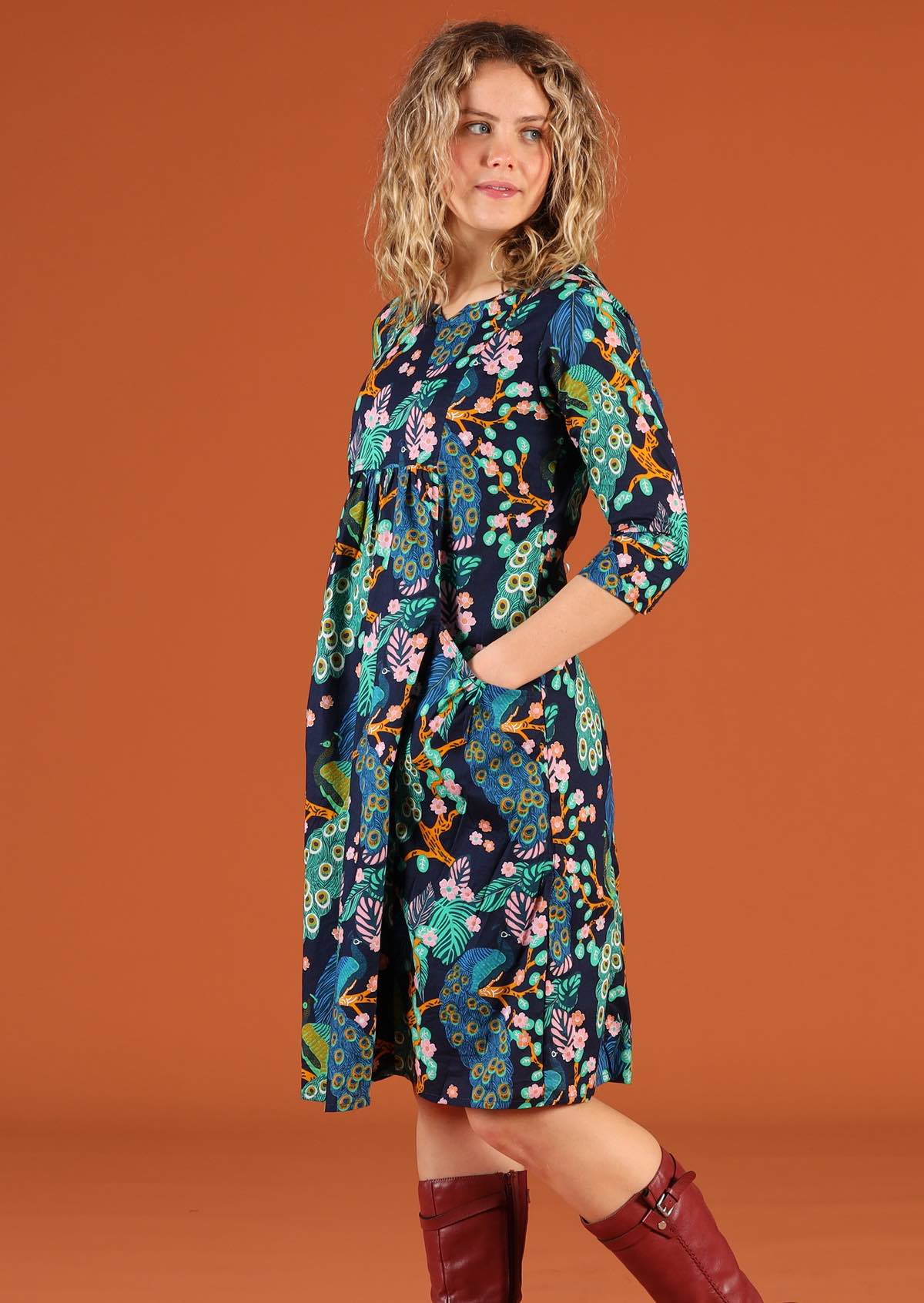 Woman standing side on wearing a knee length cotton dress in a floral peacock print with hands in pockets
