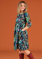 Woman standing side on wearing a knee length cotton dress in a floral peacock print with hands in pockets