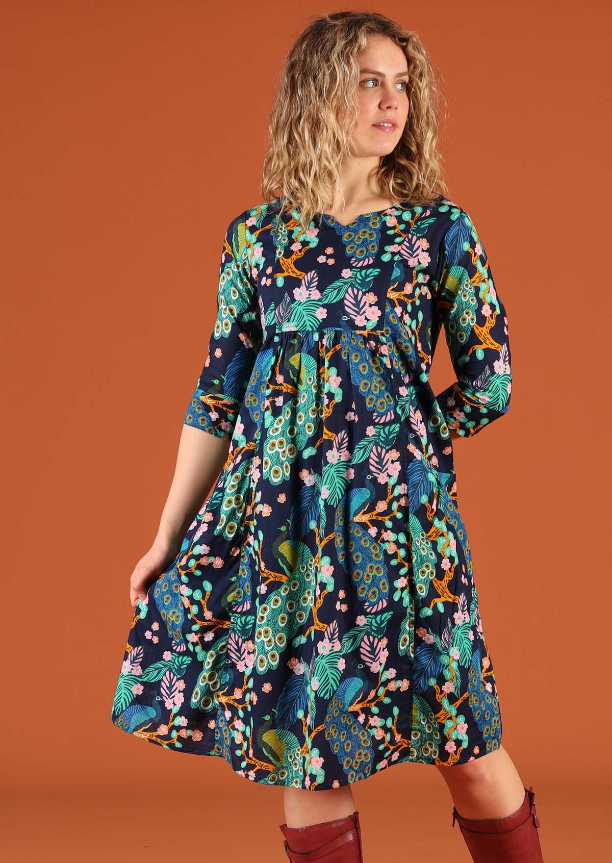 Woman wearing a knee length cotton dress in a peacock print, holding the skirt out to side