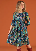 Woman wearing a knee length cotton dress in a peacock print, holding the skirt out to side