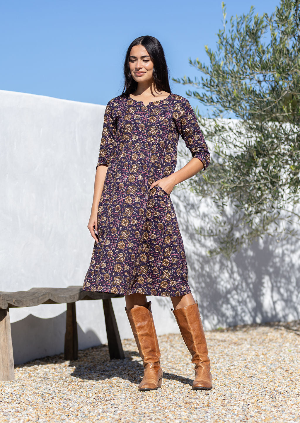 Woman wearing a purple cotton patterned dress and brown boots standing outdoors with a white wall and tree in the background.