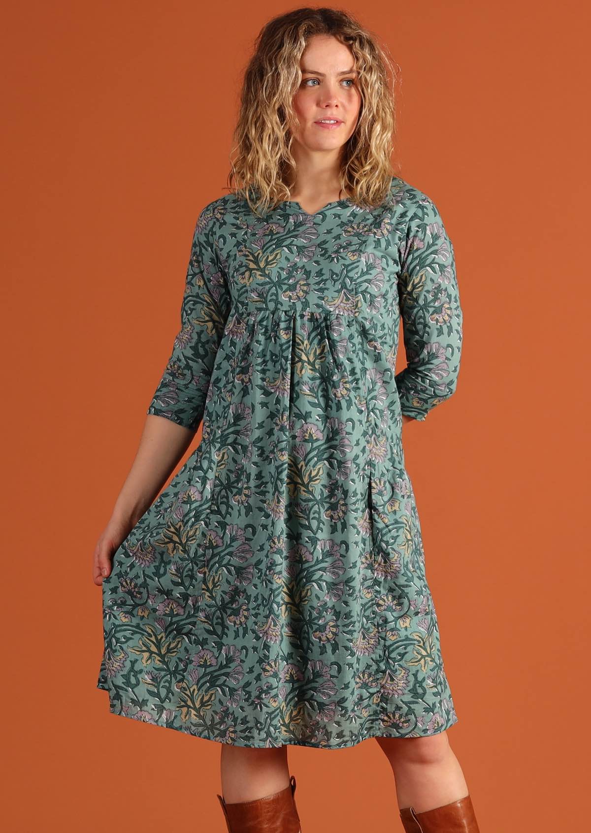  Model in front of orange background wears empire waistline relaxed fit cotton dress with pockets.