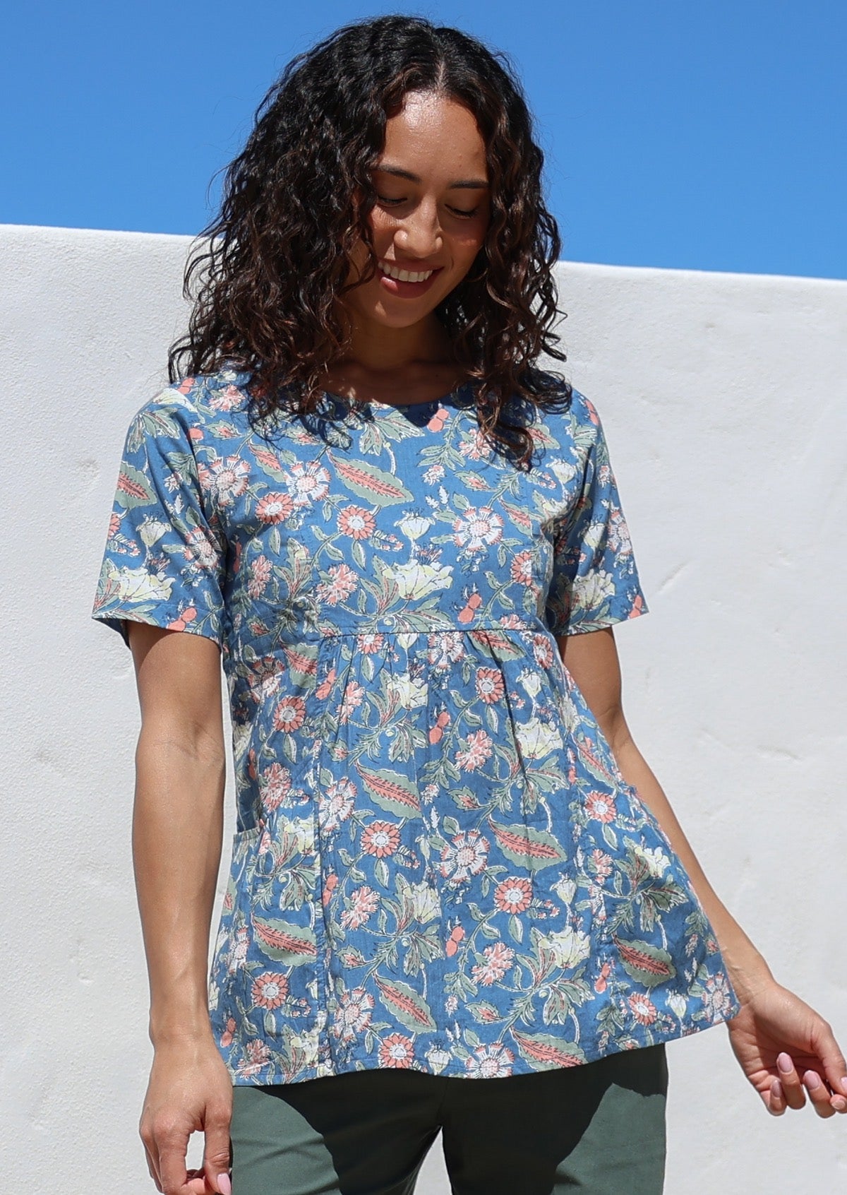 Woman wearing a dusty blue based floral print top with short sleeves and an empire waistline, with deep pockets