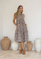 Woman in a sleeveless, floral light brown cotton dress standing in front of decorative vases with both the hands in pockets.