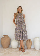 Woman standing in front of ceramic vases wears relaxed floral cotton dress with taupe base and pink, green and beige floral print.