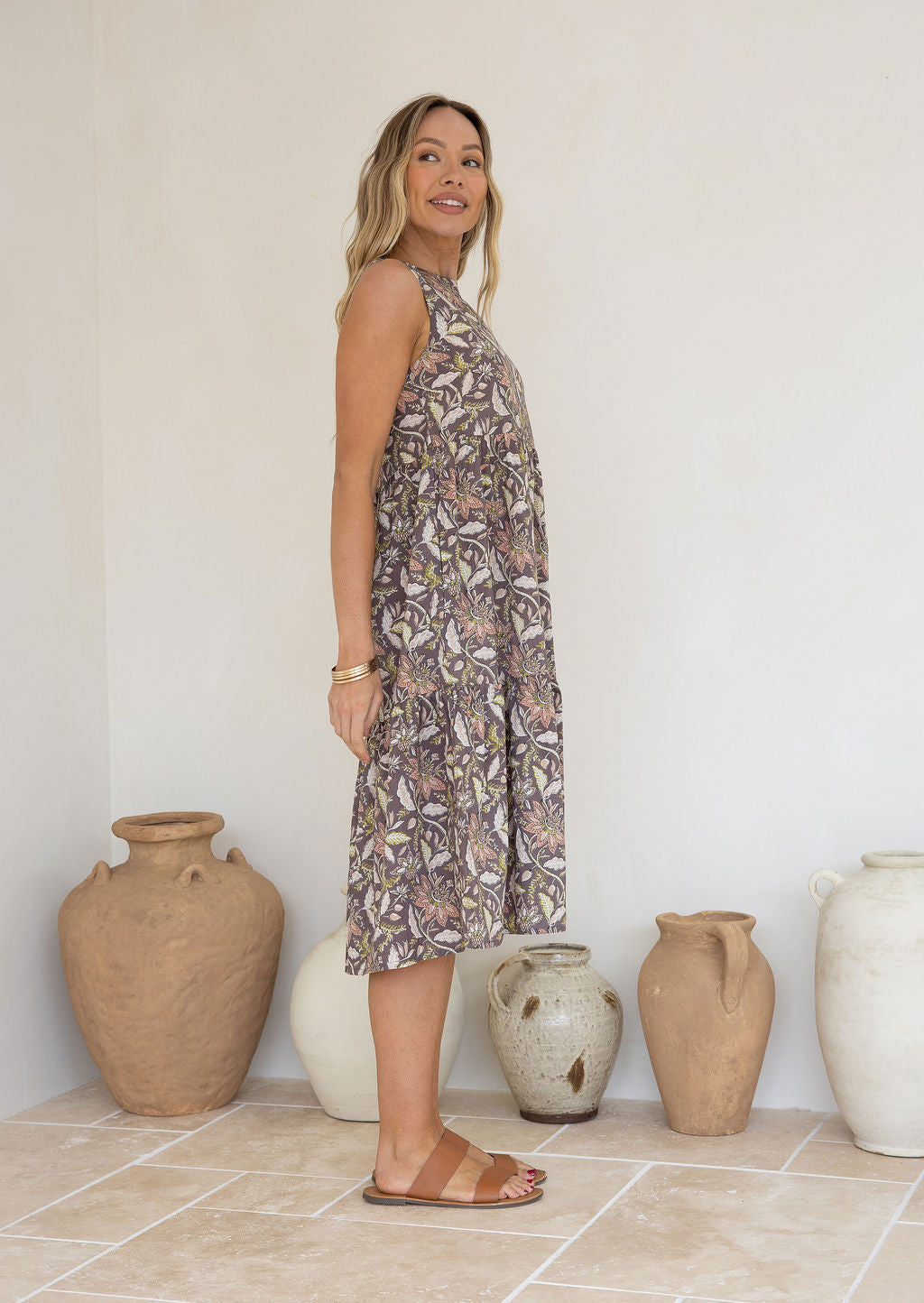 Woman stands in side pose wearing a tiered sleeveless sundress with pockets, high round neck and empire waist.
