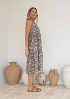 Woman stands in side pose wearing a tiered sleeveless sundress with pockets, high round neck and empire waist.
