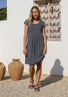 Woman in a knee length navy blue cotton  dress with cap sleeves, standing in front of a white wall with decorative elements