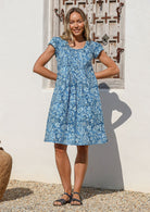 Woman wearing a blue cotton floral dress standing against a white wall with a decorative window