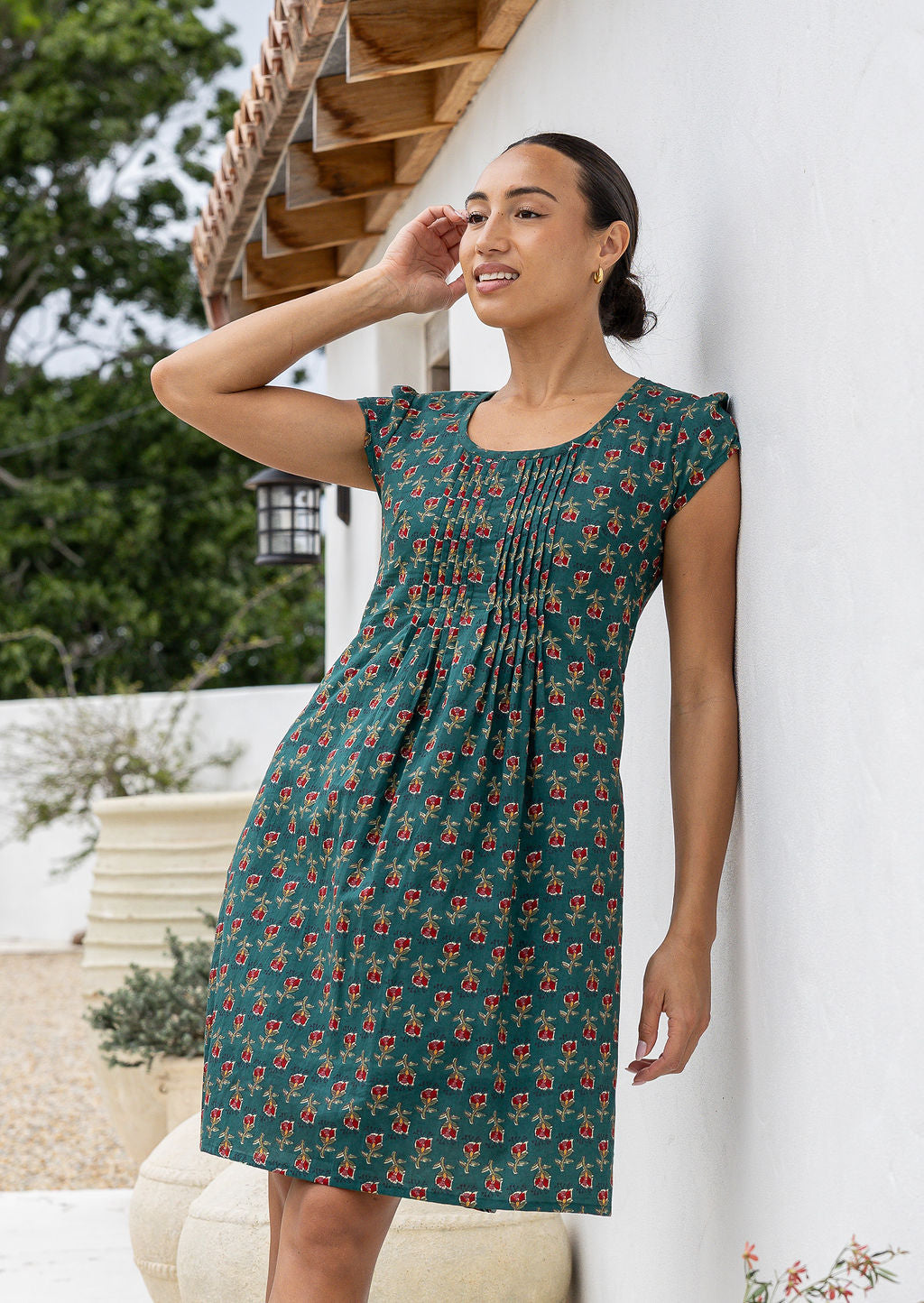 Woman wearing a Indian Cotton green floral dress standing against a white wall with trees in the background