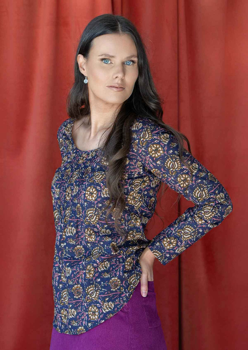 Woman standing sideways with hand in back pocket of skirt, wearing 100% cotton long sleeve top with tiny pleated details and round neck.