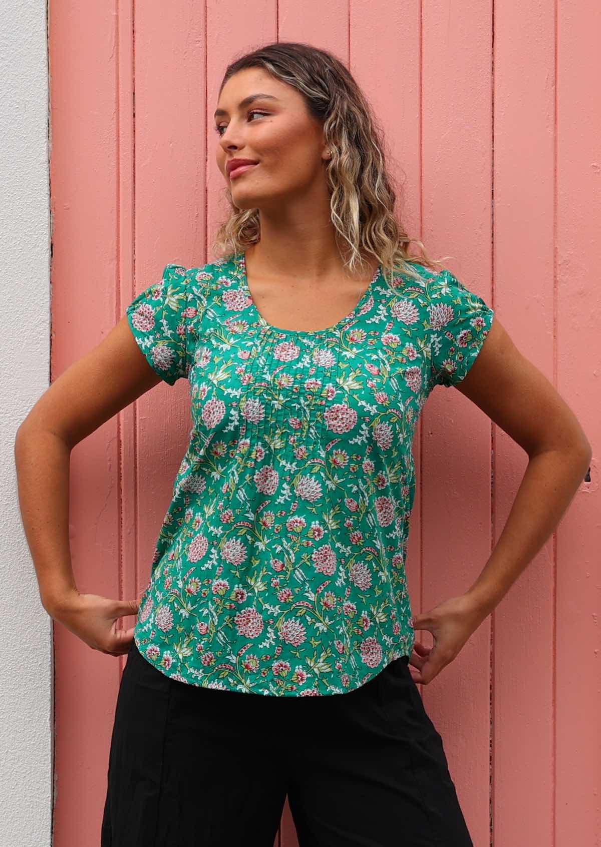 Model standing in front of pink door holding on to bottom of shirt wearing 100 percent cotton short sleeve pleated top 