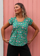 Model standing in front of pink door holding on to bottom of shirt wearing 100 percent cotton short sleeve pleated top 