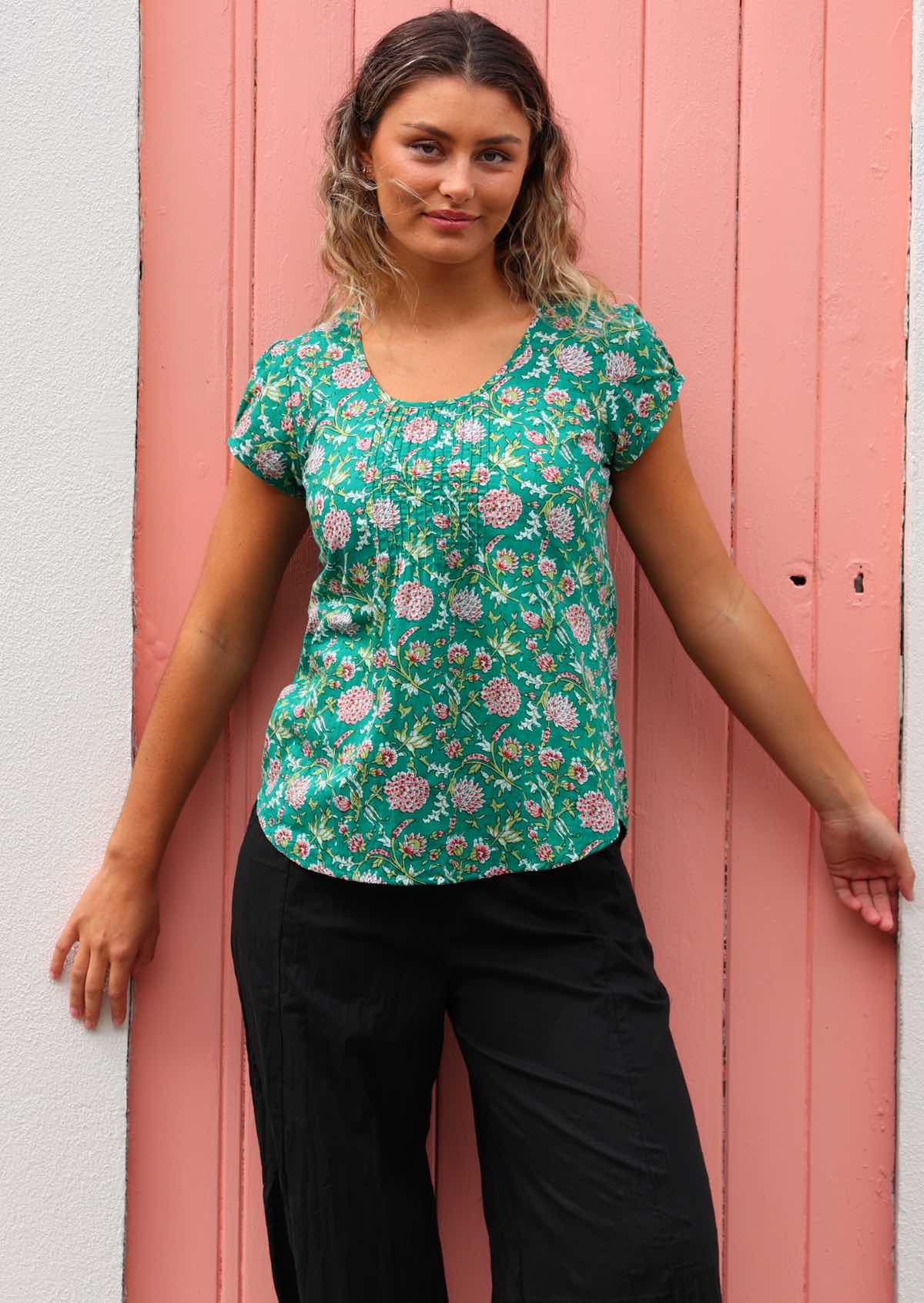 Model standing in front of pink door wearing 100 percent cotton short sleeve pleated top paired with black pants