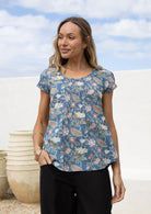 Woman wears dusty blue based floral cotton cap sleeve top with pin tuck detail across the bust and a scoop neckline