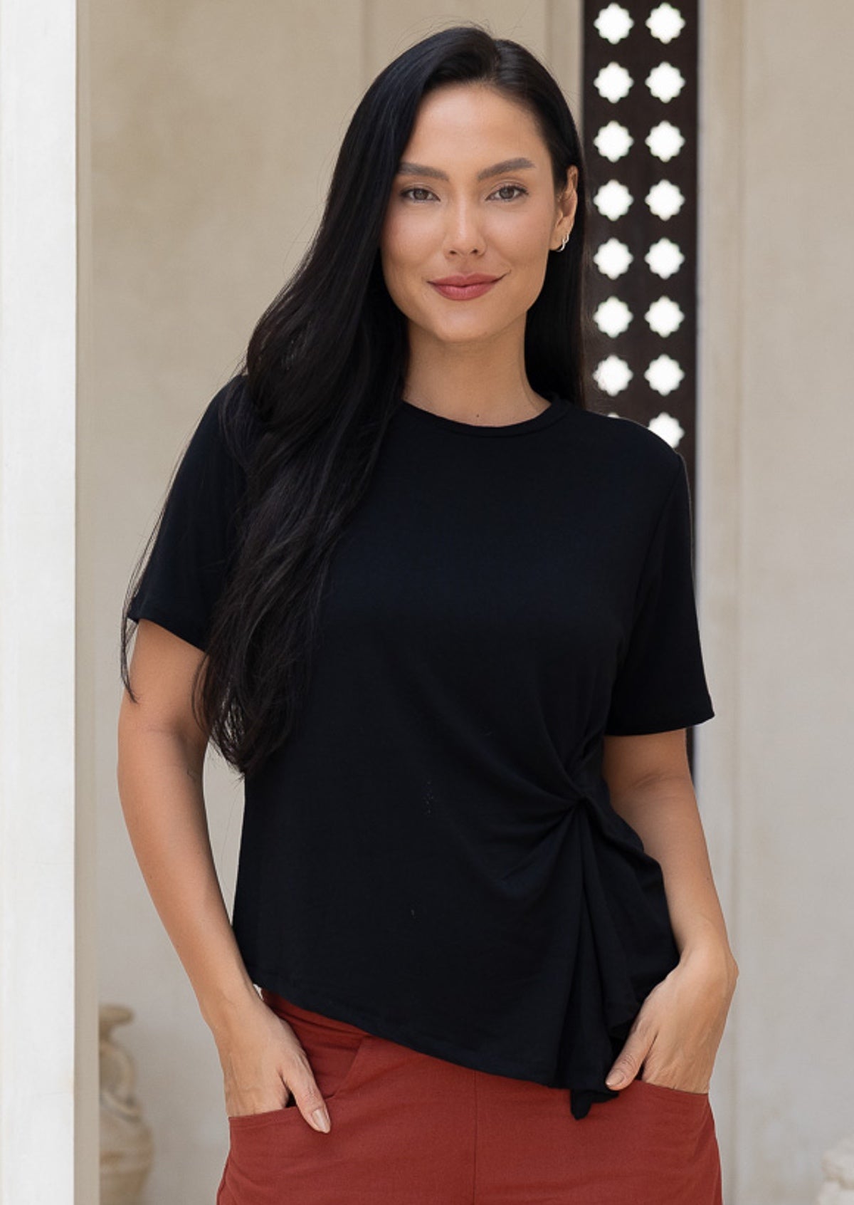 Woman wearing a black top with a knot detail with hands in pockets of rust pants