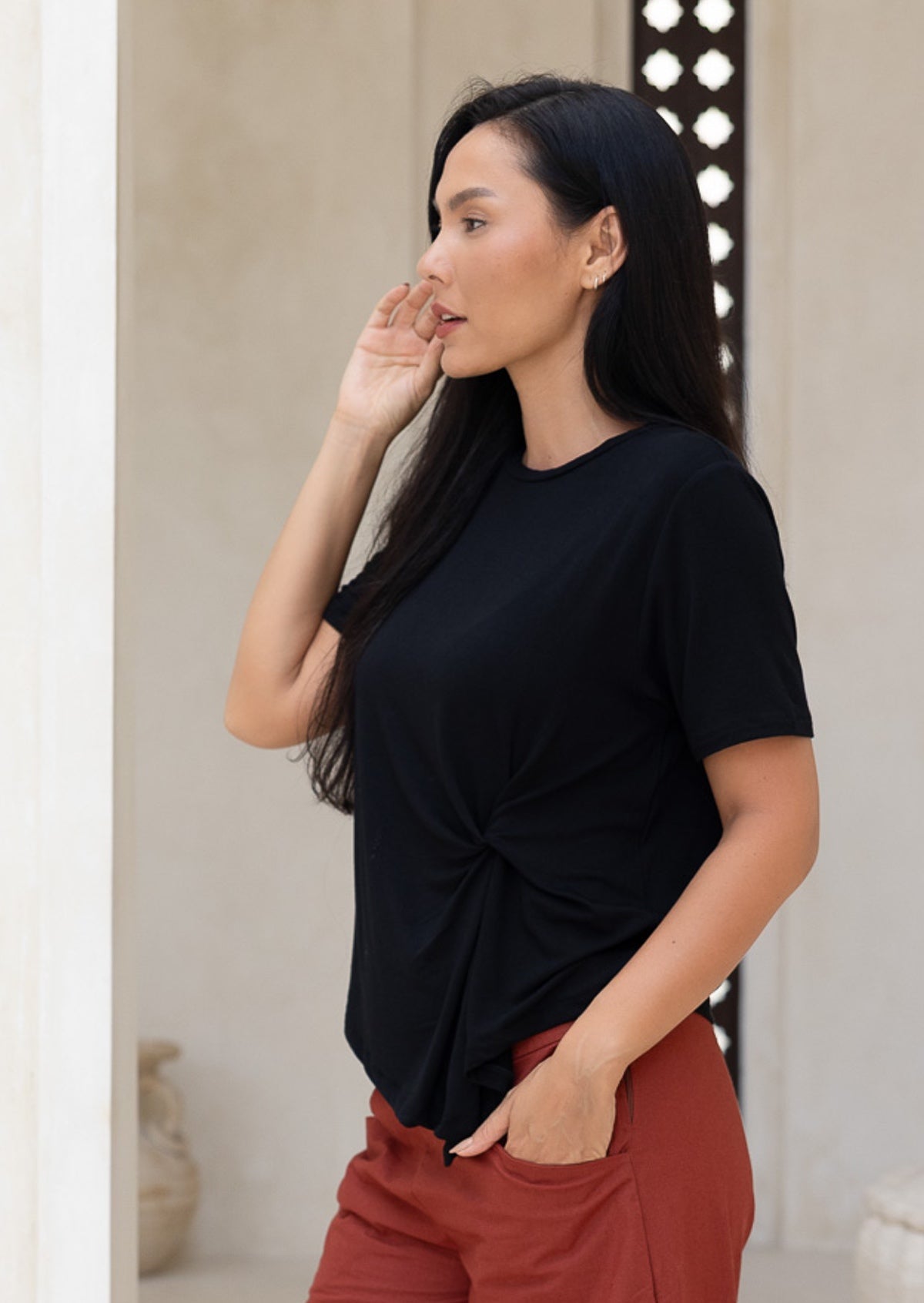 Woman wearing a black top and rust pants standing side on with a hand in pocket