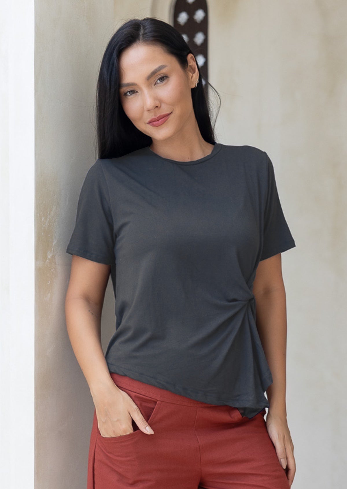Woman wears dark grey rayon t-shirt with short sleeves and knot detail on the front left, paired with rust pants with hand in pocket