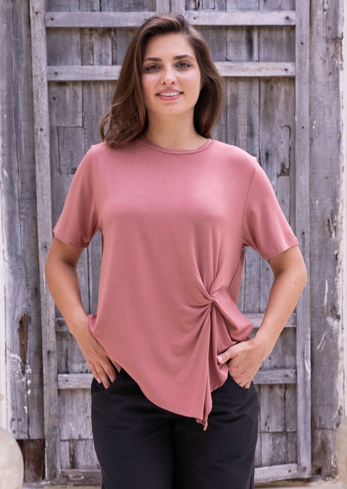 Woman wearing a pink t-shirt with a knot detail over black cotton pants in front of a wooden fence.