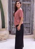 Woman wearing a loose fit pink t-shirt with a knot detail over black cotton pants in front of a wooden fence.