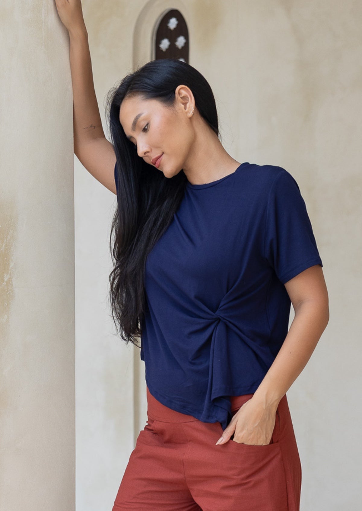 Woman wearing a navy blue stretch rayon top with a knot detail and rust pants against a neutral wall.
