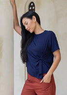 Woman wearing a navy blue stretch rayon top with a knot detail and rust pants against a neutral wall.