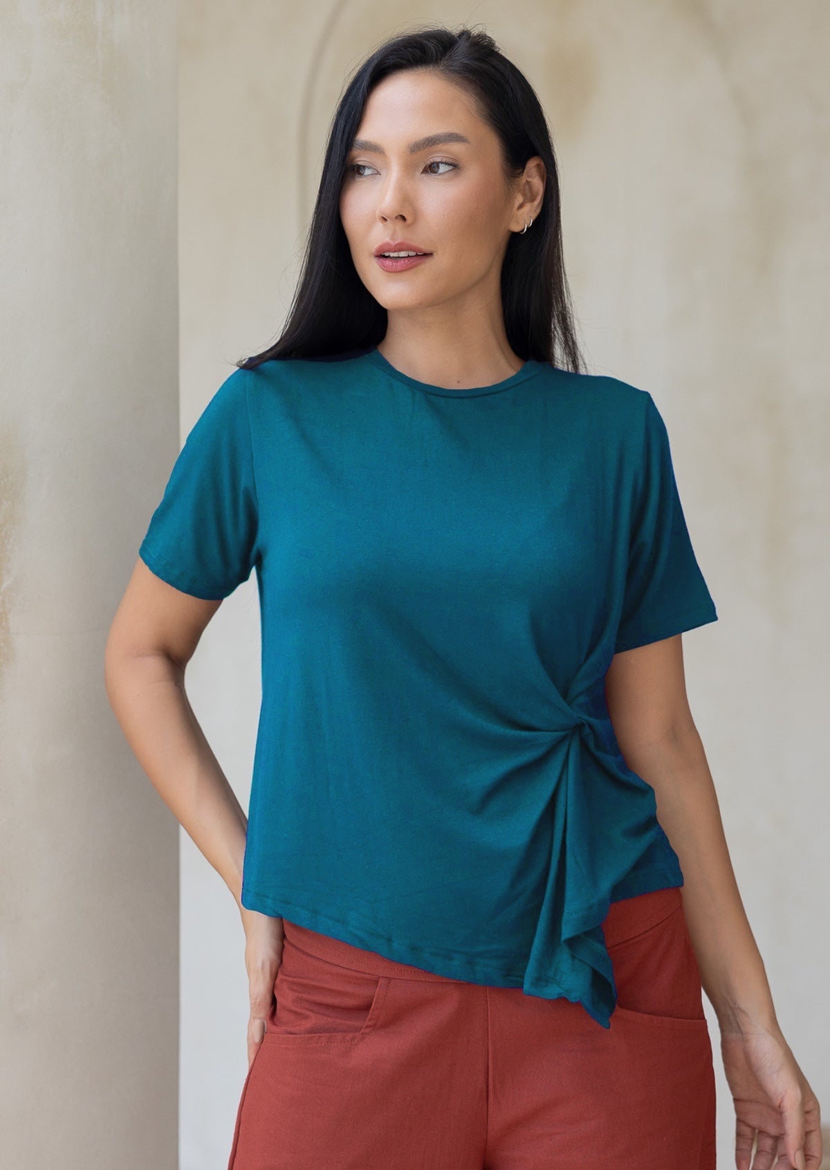 Woman wearing a teal rayon t-shirt with a knot detail, standing in front of a white wall.