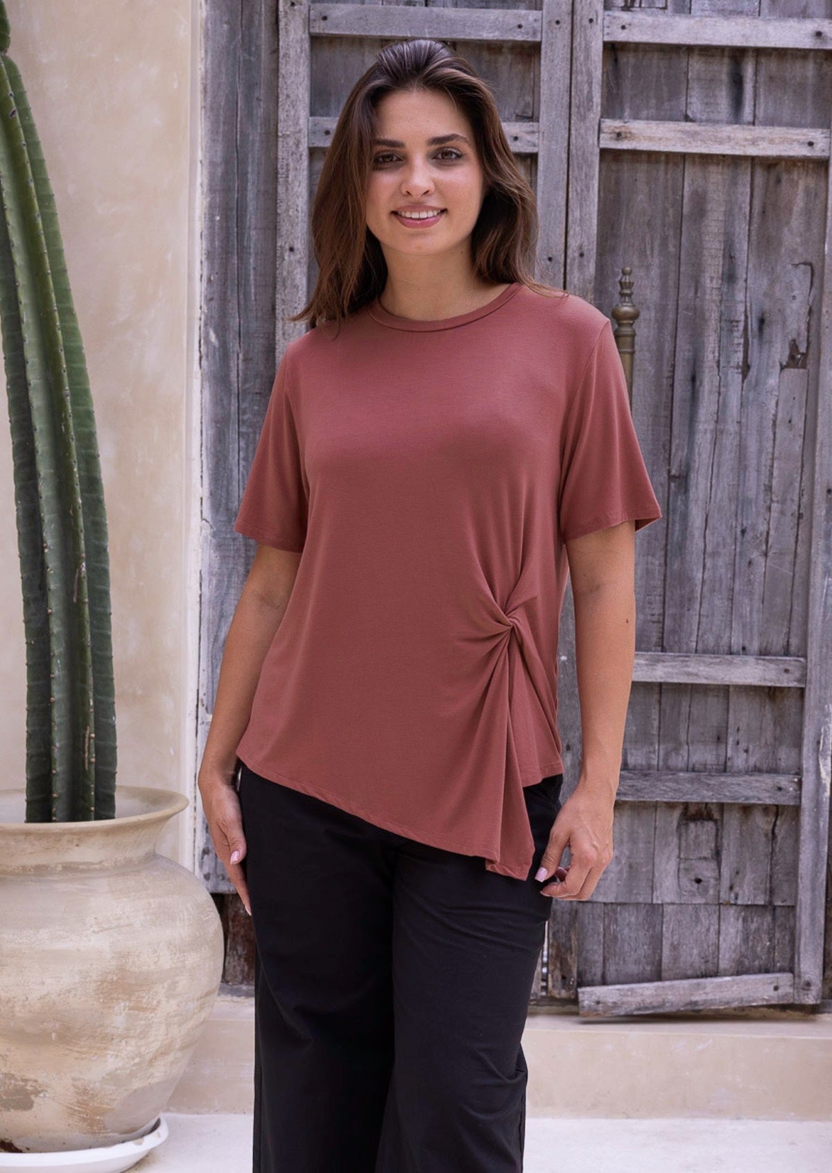 Woman wearing a terracotta short sleeved rayon top with a knot detail, standing against a wooden door