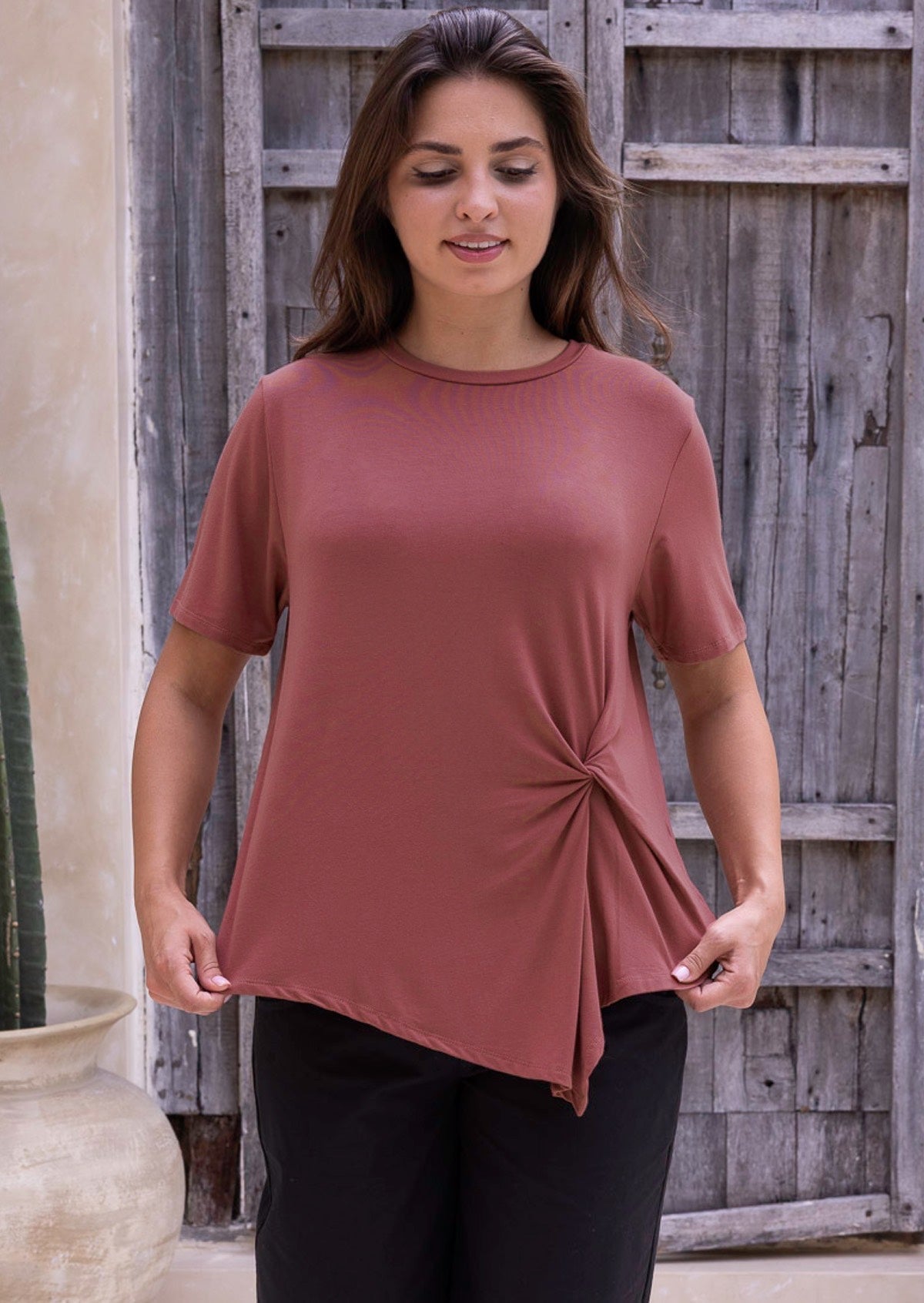 Woman wearing a terracotta coloured top with a knot detail, standing outdoors next to a wooden fence and cactus.