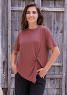 Woman wearing a terracotta rayon top with a knot detail in the front, standing against a wooden wall.