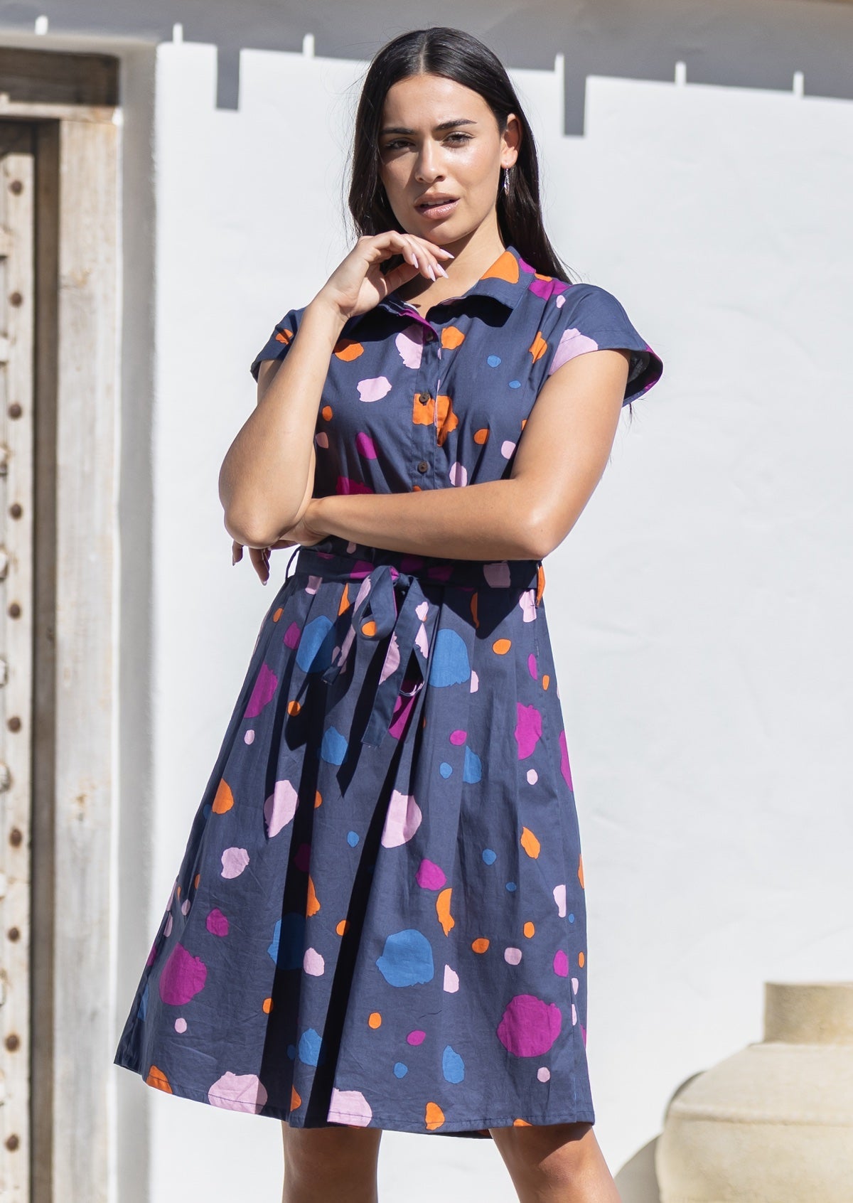 Woman wearing a colourful cotton shirt dress with abstract patterns against a white wall.