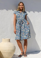 Woman in a blue floral cotton dress with cap sleeves, a buttoned bodice, pockets, box pleated at the waist and a fabric waist tie.