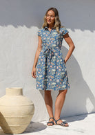 Model stands with one hand in pocket wearing a 100% cotton knee length shirt style retro floral dress in dusty blue