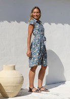 Woman in a blue floral shirt style cotton dress with cap sleeves, pockets, box pleated at the waist and a fabric waist tie.