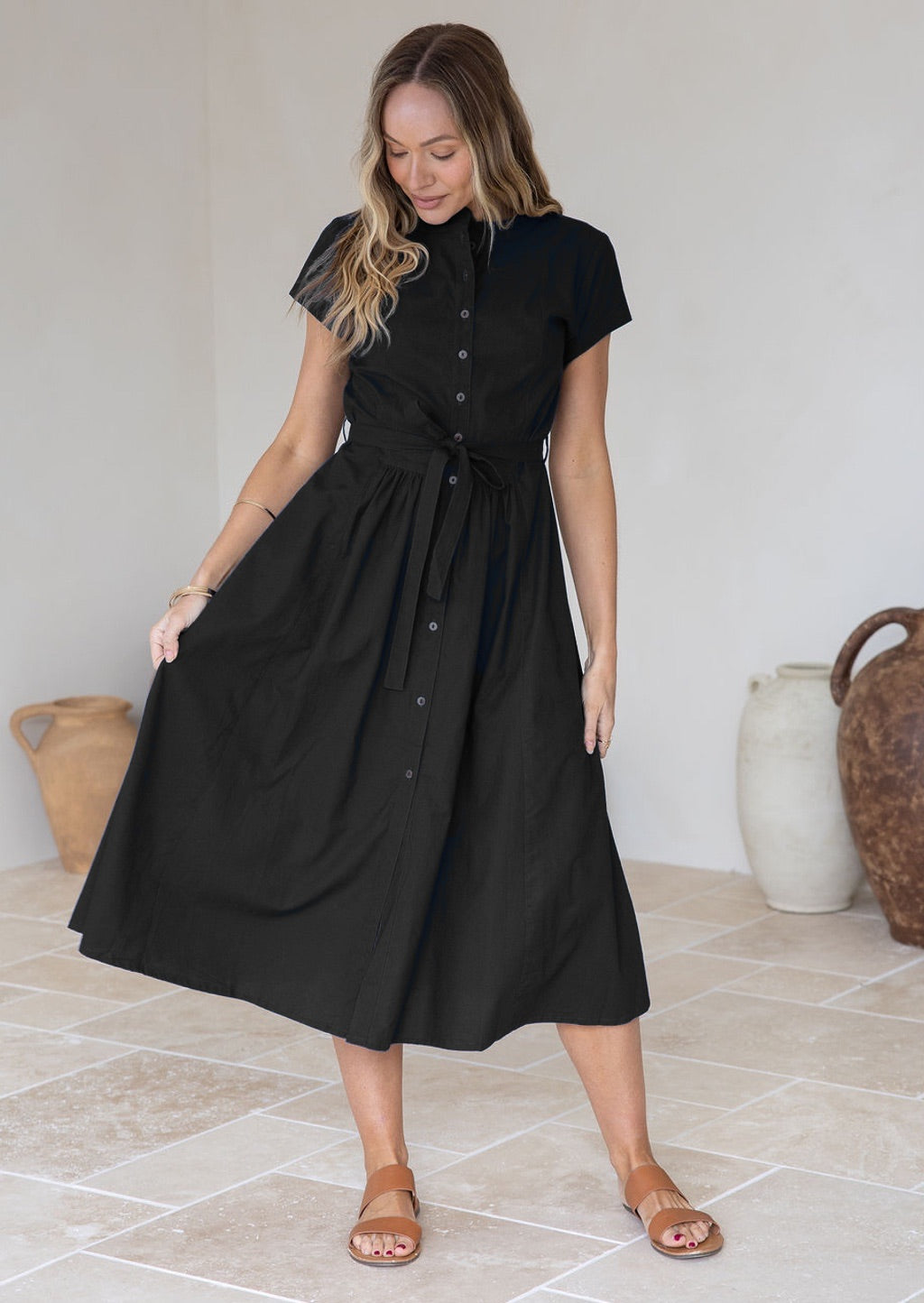 Woman wearing a 50's style black cotton button through dress, holding the skirt out to the side