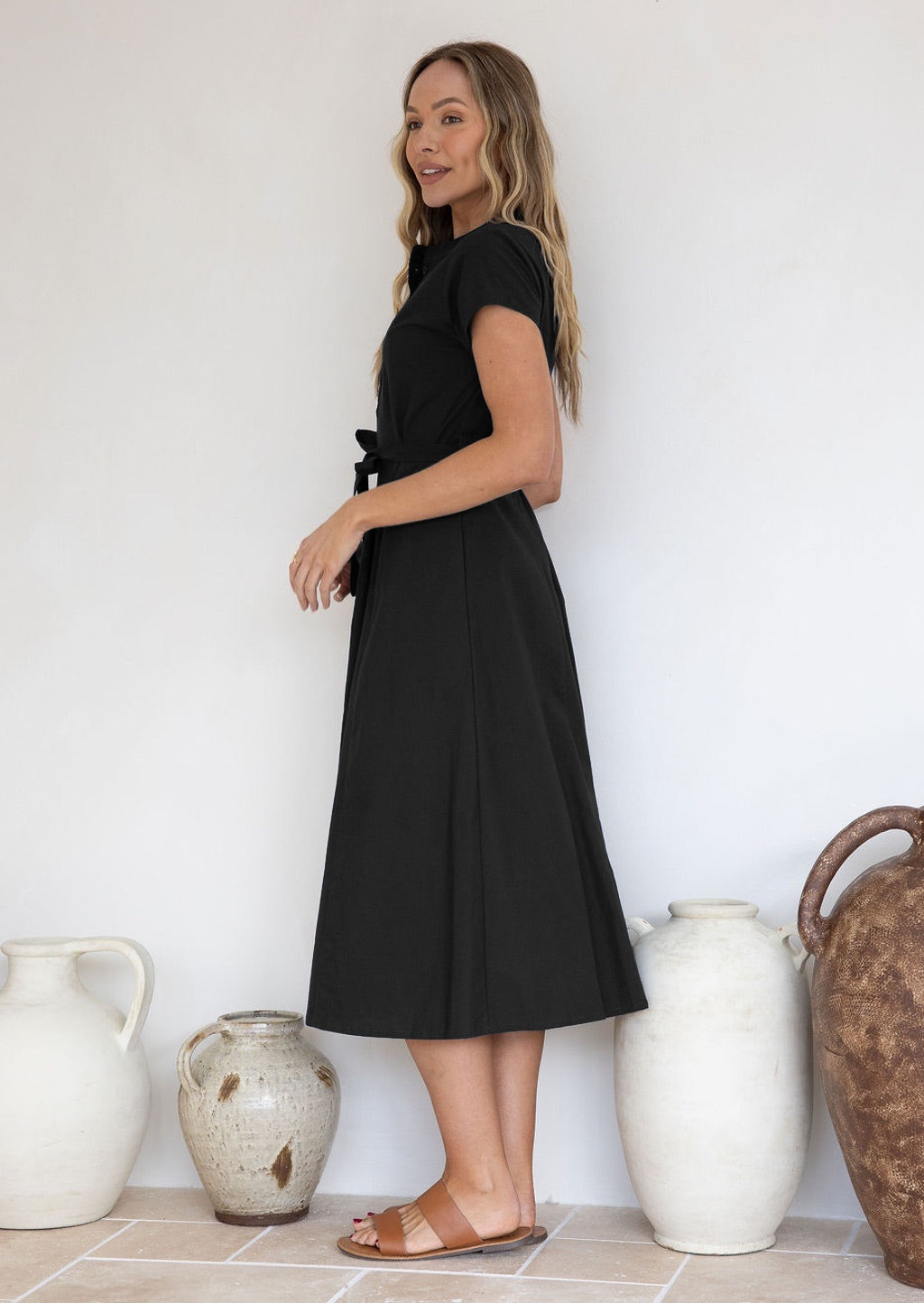 Woman wearing a black 50's style midi length dress with cap sleeves, standing in front of decorative vases.