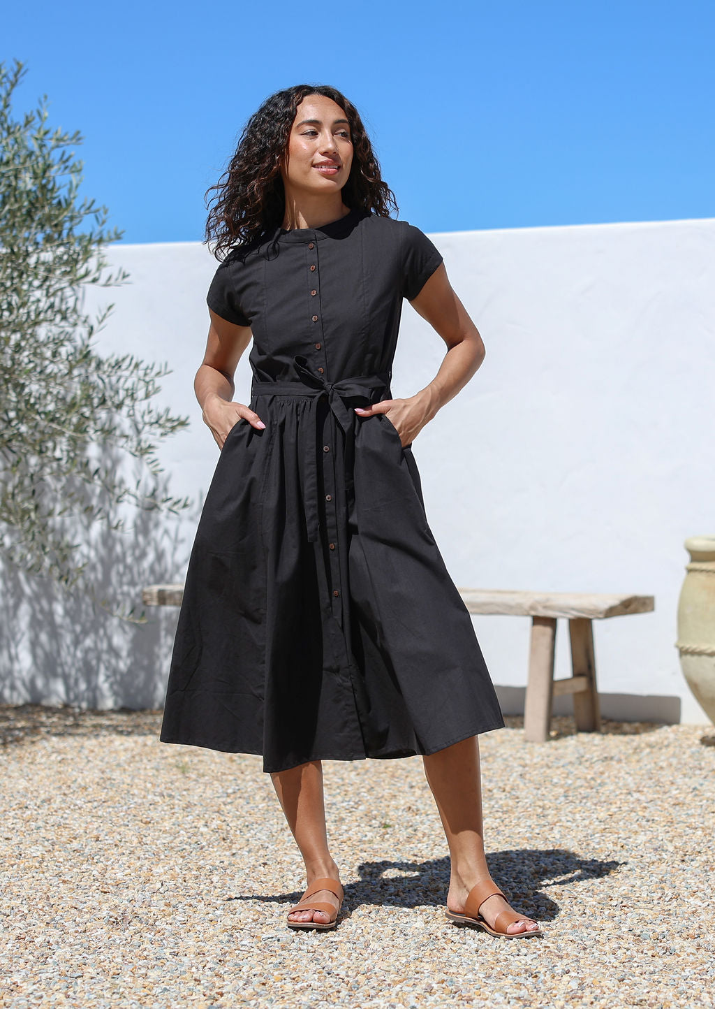 Woman wearing a black cotton retro style dress standing outdoors with a white wall with hands in pockets.