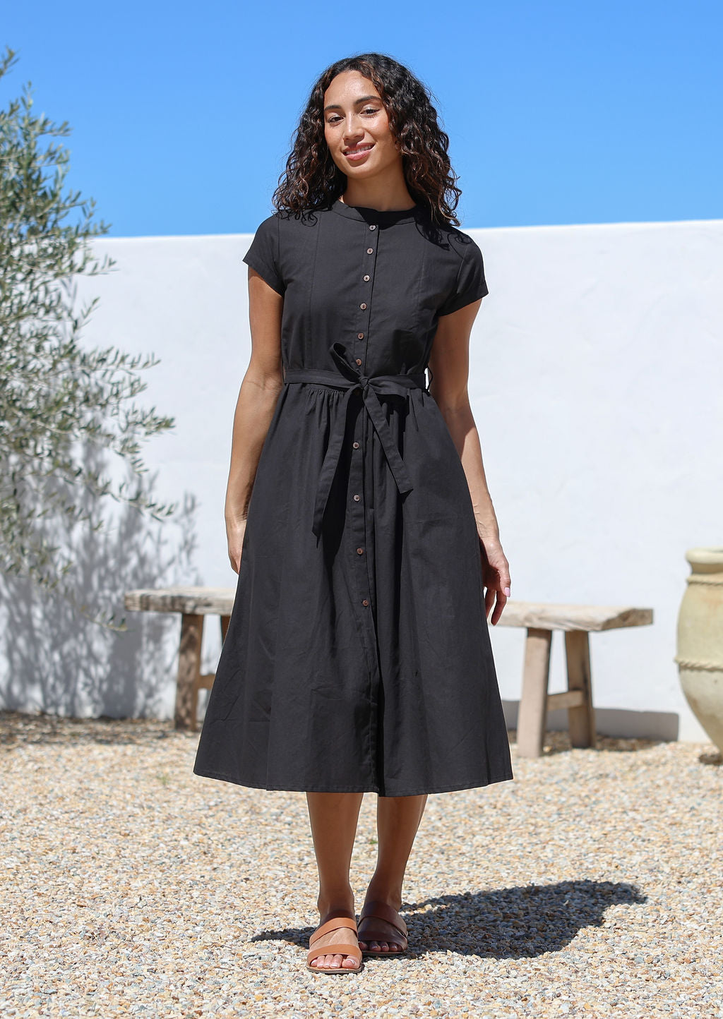 Woman wearing a black cotton retro style dress with button down front standing outdoors with a white wall with hands in pockets.