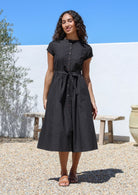 Woman wearing a black cotton retro style dress with button down front standing outdoors with a white wall with hands in pockets.
