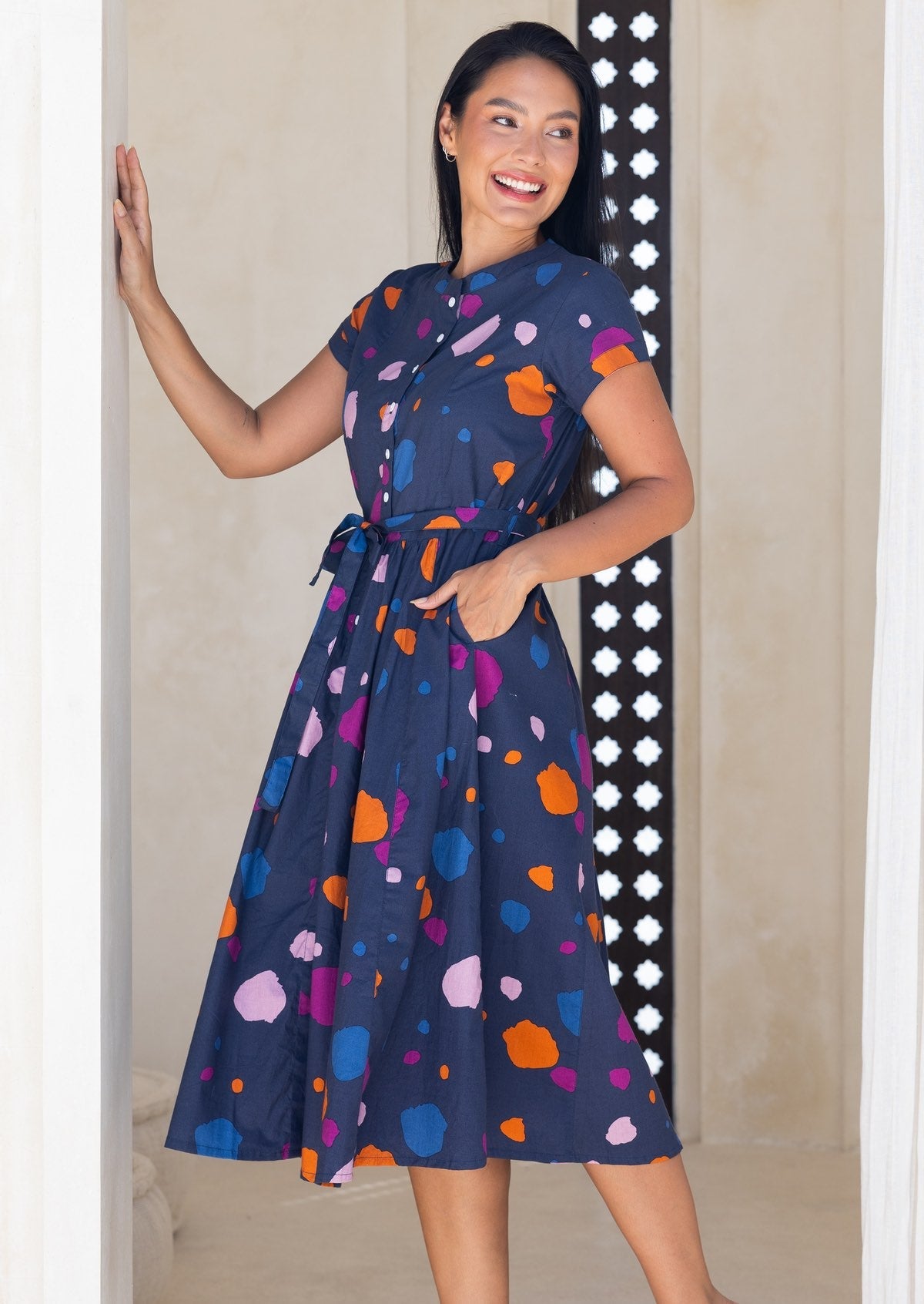 Woman wearing a navy based cotton 50's style dress with colourful polka dot print, with hand in side pocket and waist tie