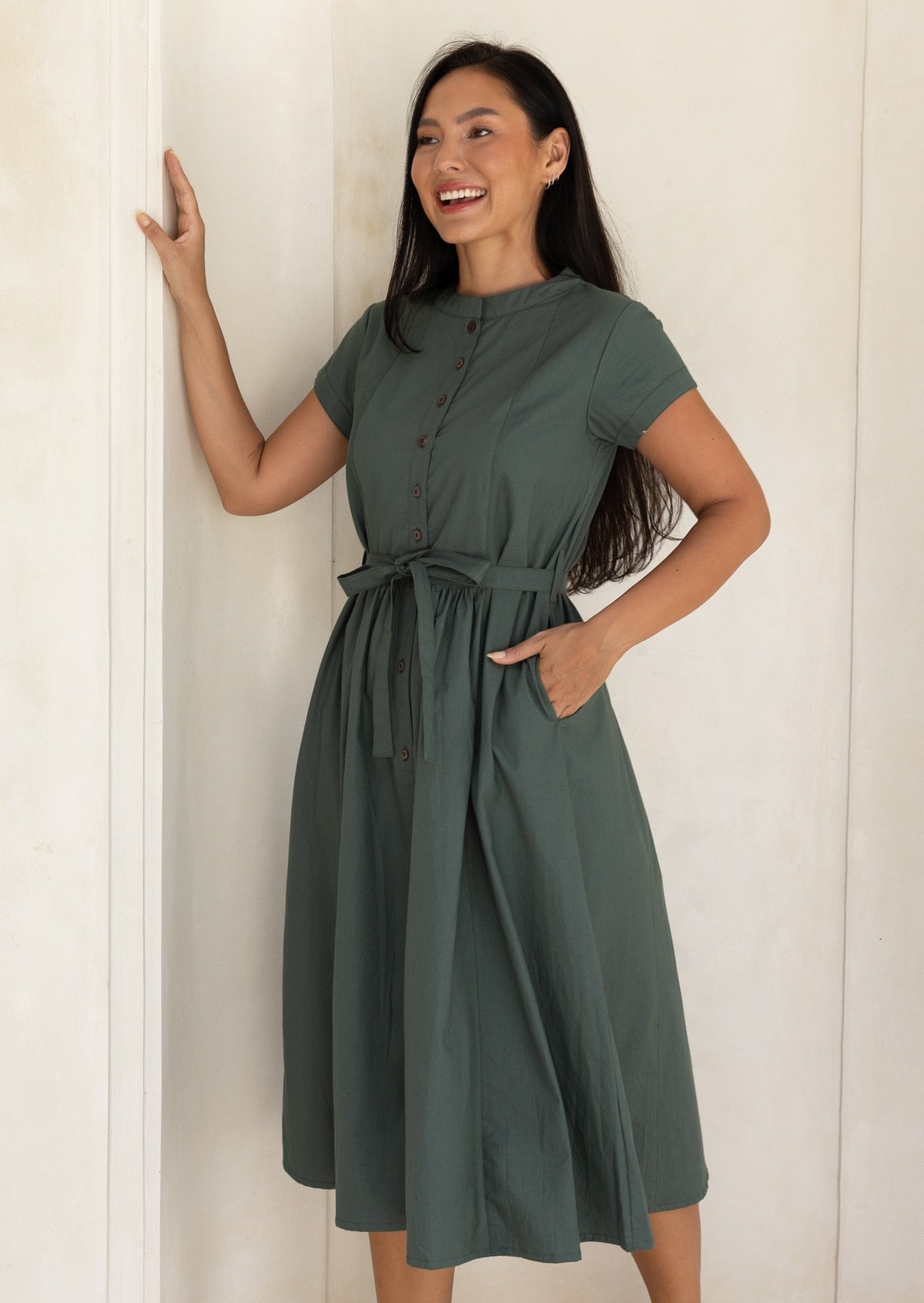 Woman wearing a moss green cotton dress standing against a white wall, with hand in side pocket