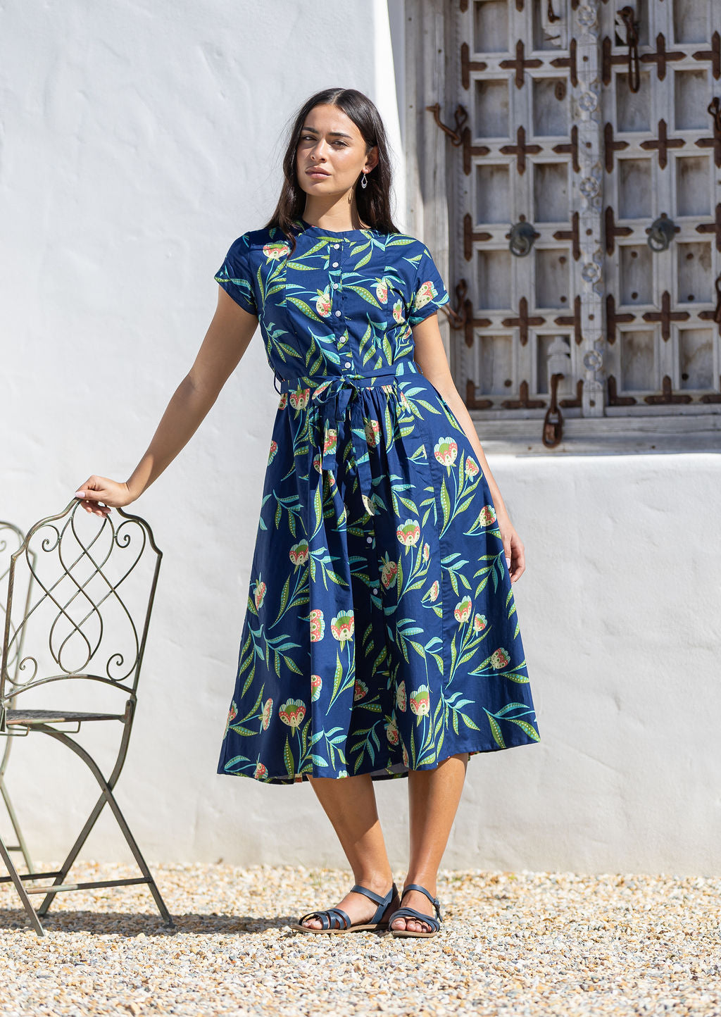 Woman wearing a blue floral cotton midi dress standing next to a chair against a white wall.