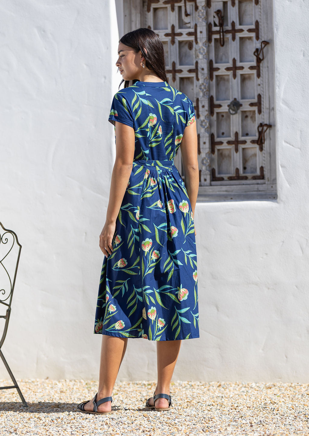 Woman wearing a blue floral cotton sundress standing in front of a white wall with a decorative metal gate.