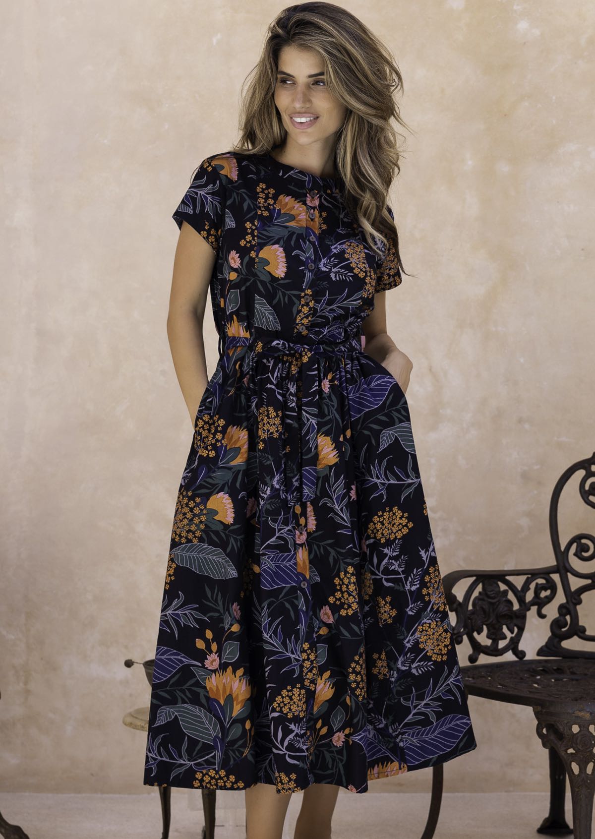 Model wearing a black floral cotton, button through dress with removable waist tie, short sleeves and hands in pocket.