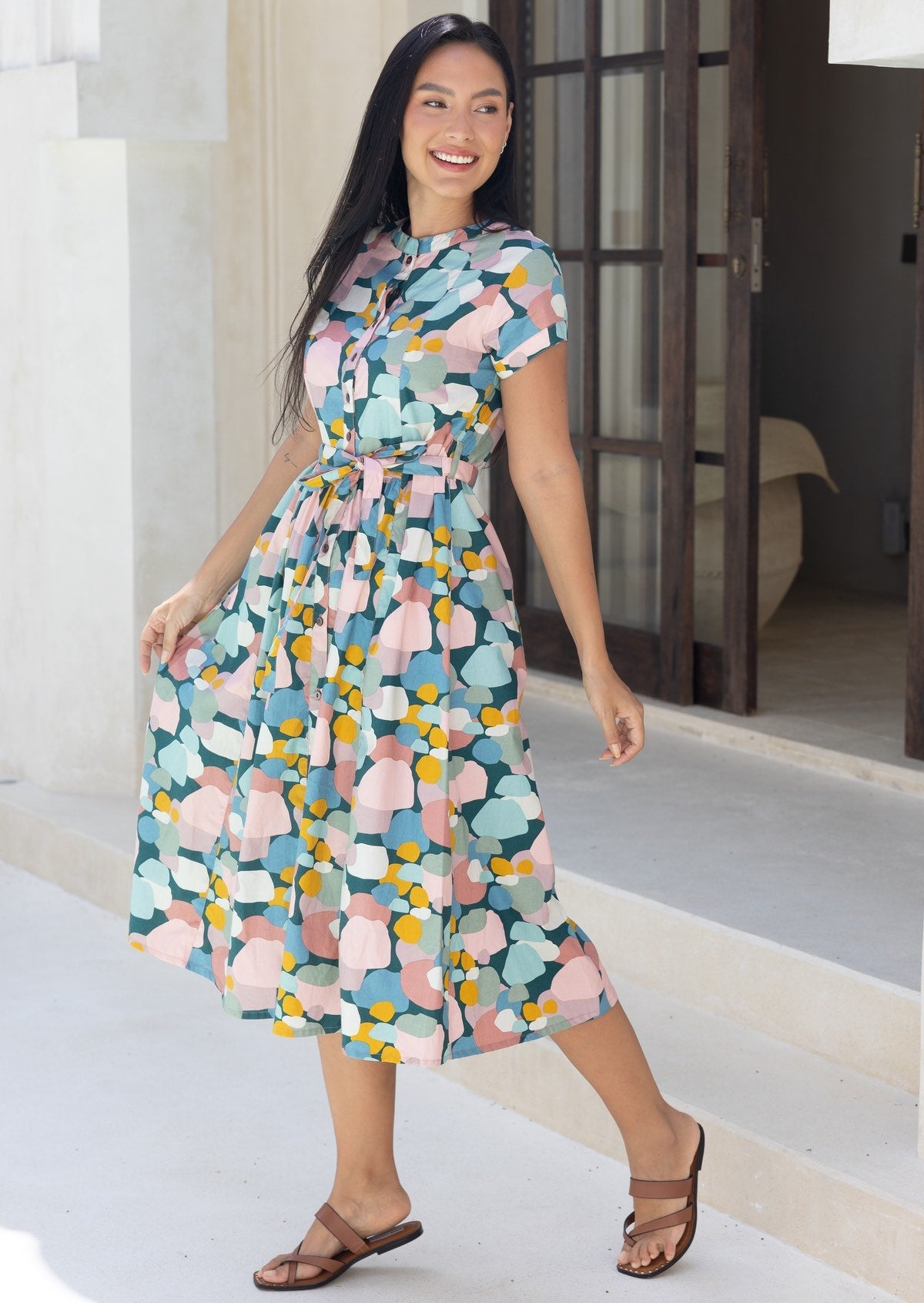 Woman standing sideways in cotton 50's style midi length dress in pastel colours with waist tie