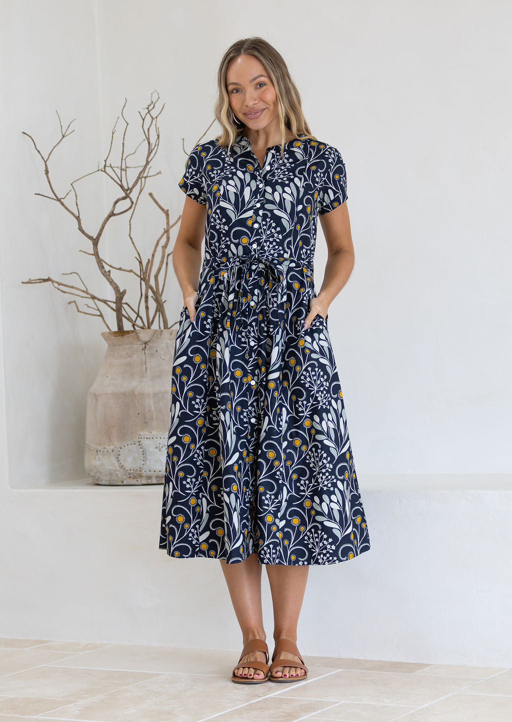 Woman wearing a black floral patterned cotton dress standing in a minimalistic room with a vase and branches.
