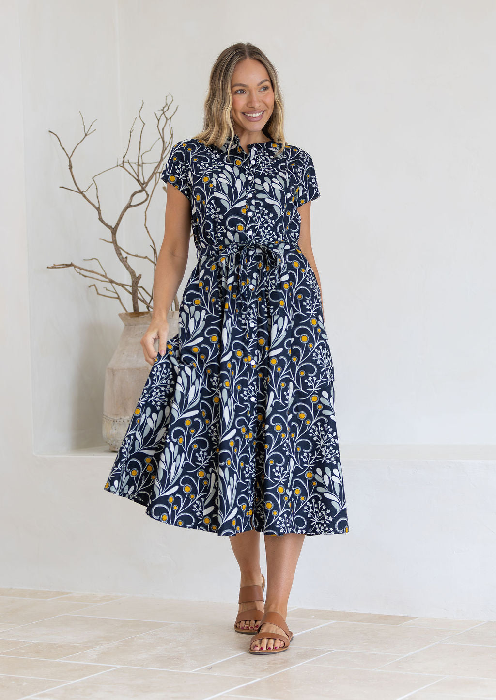 Woman wearing a dark cotton floral dress standing in a minimalistic room with a vase and branches.